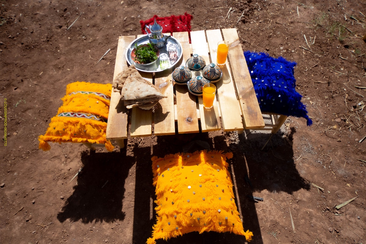 A low wooden table is surrounded by colorful cushions in shades of orange, blue, and red. Traditional Moroccan tea glasses and a decorative platter are displayed atop the table, accompanied by fresh herbs and two glasses of orange drink, enhancing the inviting setting.