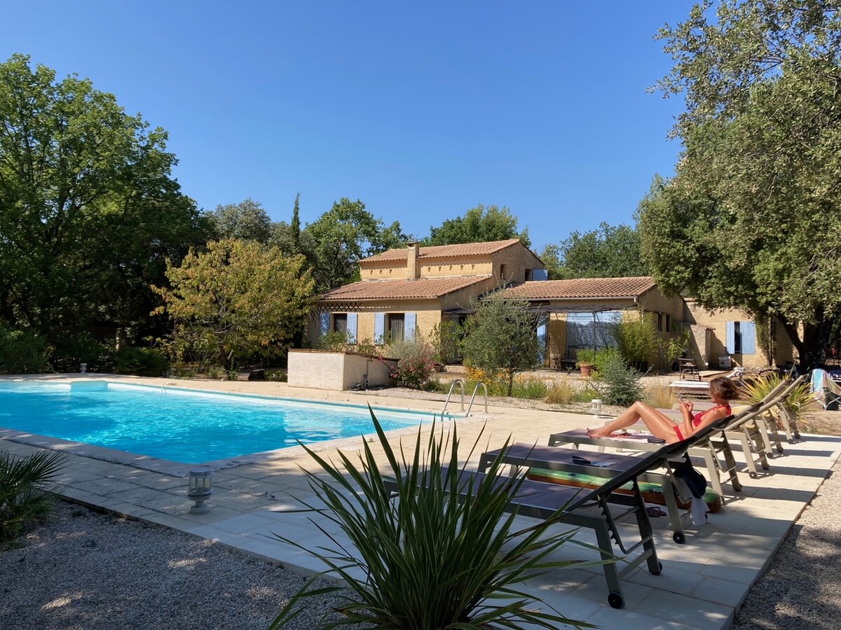 A spacious outdoor area features a large swimming pool surrounded by sun loungers. Lush trees and shrubs frame the house, which showcases multiple windows. The clear blue sky enhances the tranquil atmosphere of the setting.