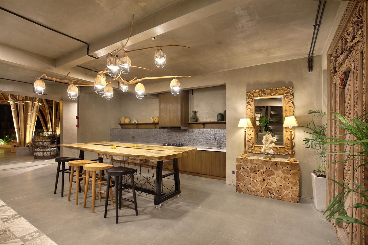 A modern kitchen space features a large wooden dining table with four stools, illuminated by an artistic hanging pendant light fixture. Warm wood cabinetry complements the neutral walls, while decorative elements and a carved mirror add a touch of character. Natural light enhances the inviting atmosphere.