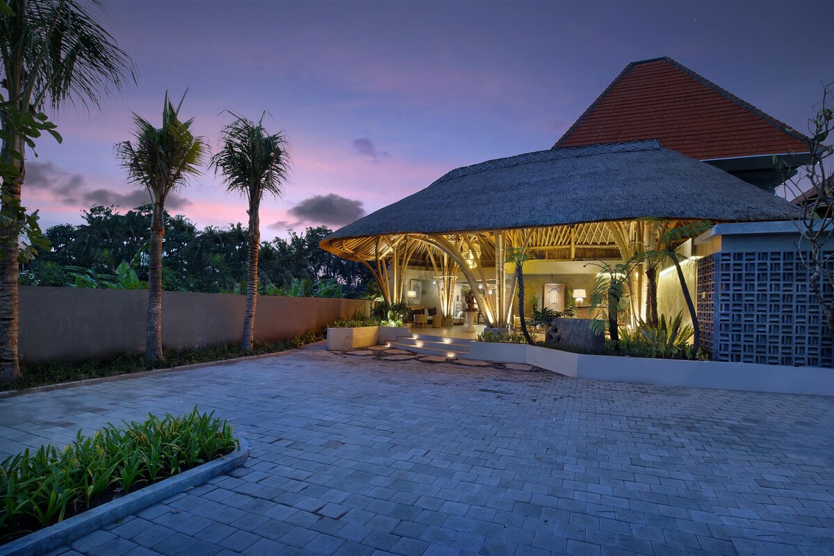 An inviting resort entrance features traditional bamboo architecture with a thatched roof, illuminated softly at dusk. Surrounding palm trees and well-maintained landscaping create a serene environment, while a spacious stone driveway leads to the entrance, enhancing accessibility and ease of arrival.