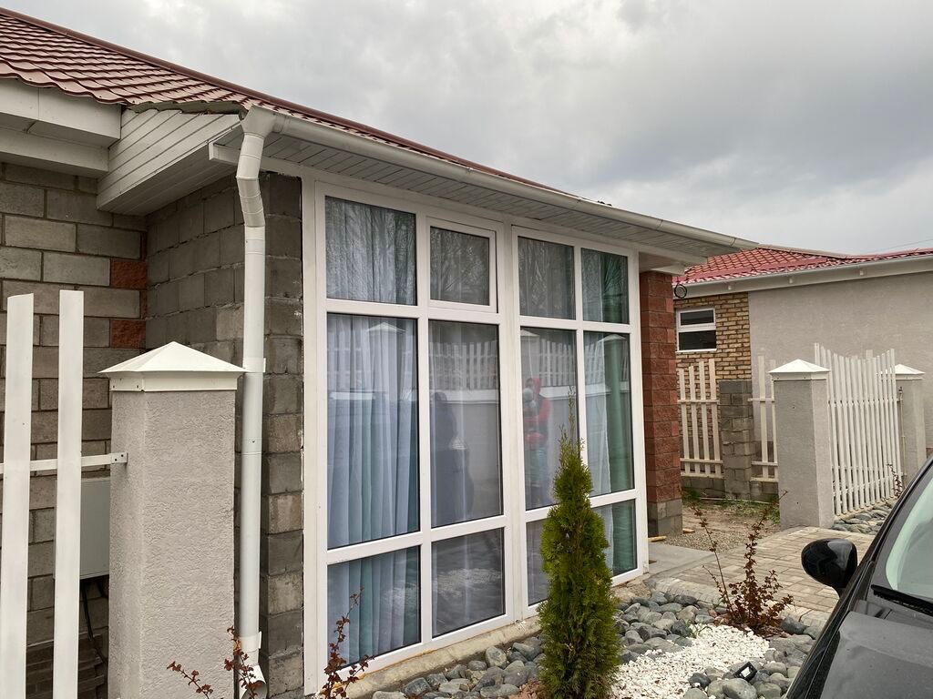 A modern cottage exterior features large windows with soft curtains, allowing natural light to flow inside. The structure is surrounded by landscaped stones and shrubbery. A driveway with vehicle parking is visible beside the cottage, indicating accessibility.