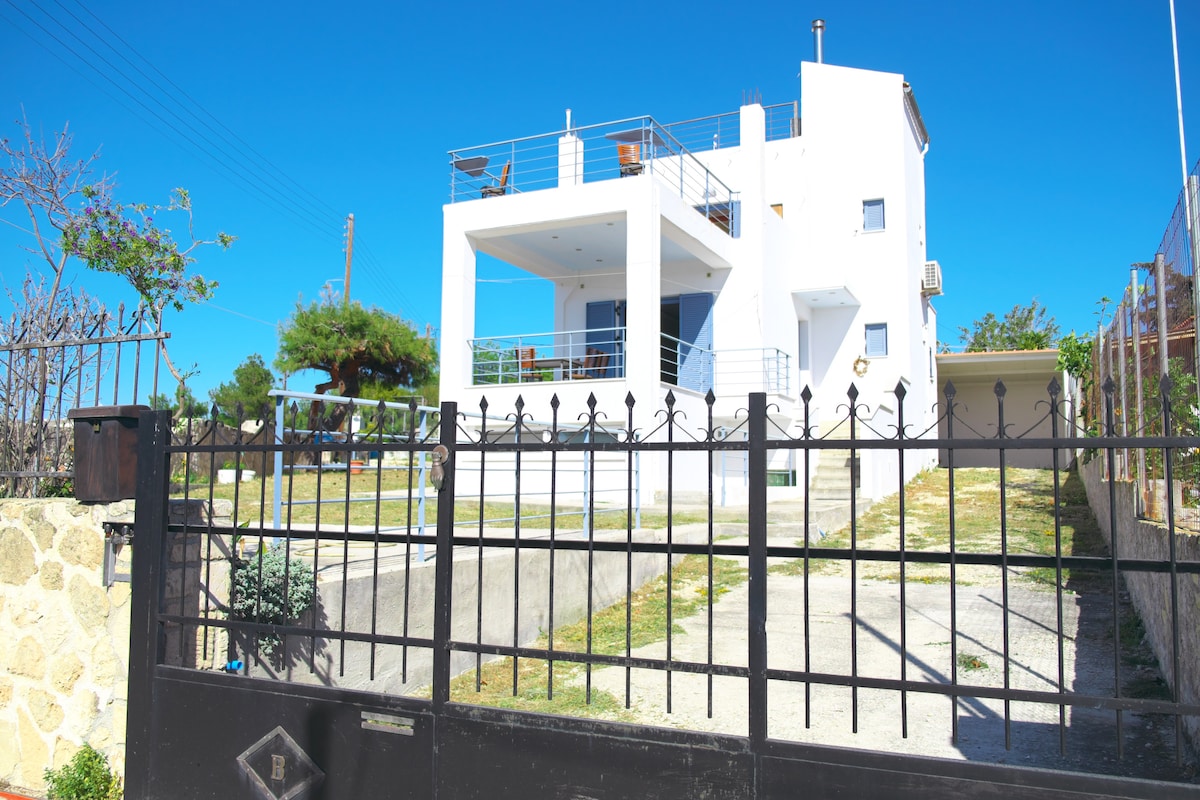 A modern, two-story house with a minimalist design stands proudly behind a gated entrance. The white exterior contrasts against the blue sky, and a spacious balcony overlooks the surrounding landscape. A driveway leads up to the house, complemented by a manicured front yard.