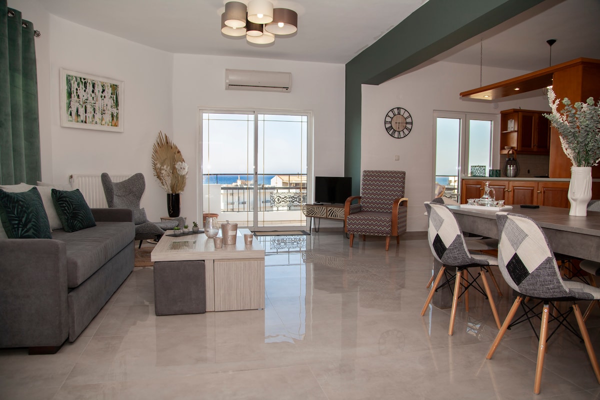A modern living area features a comfortable gray sofa and a stylish dining table surrounded by various chairs. Large windows invite natural light, offering views of the sea. The floor is glossy and light-colored, enhancing the spacious feel of the room.