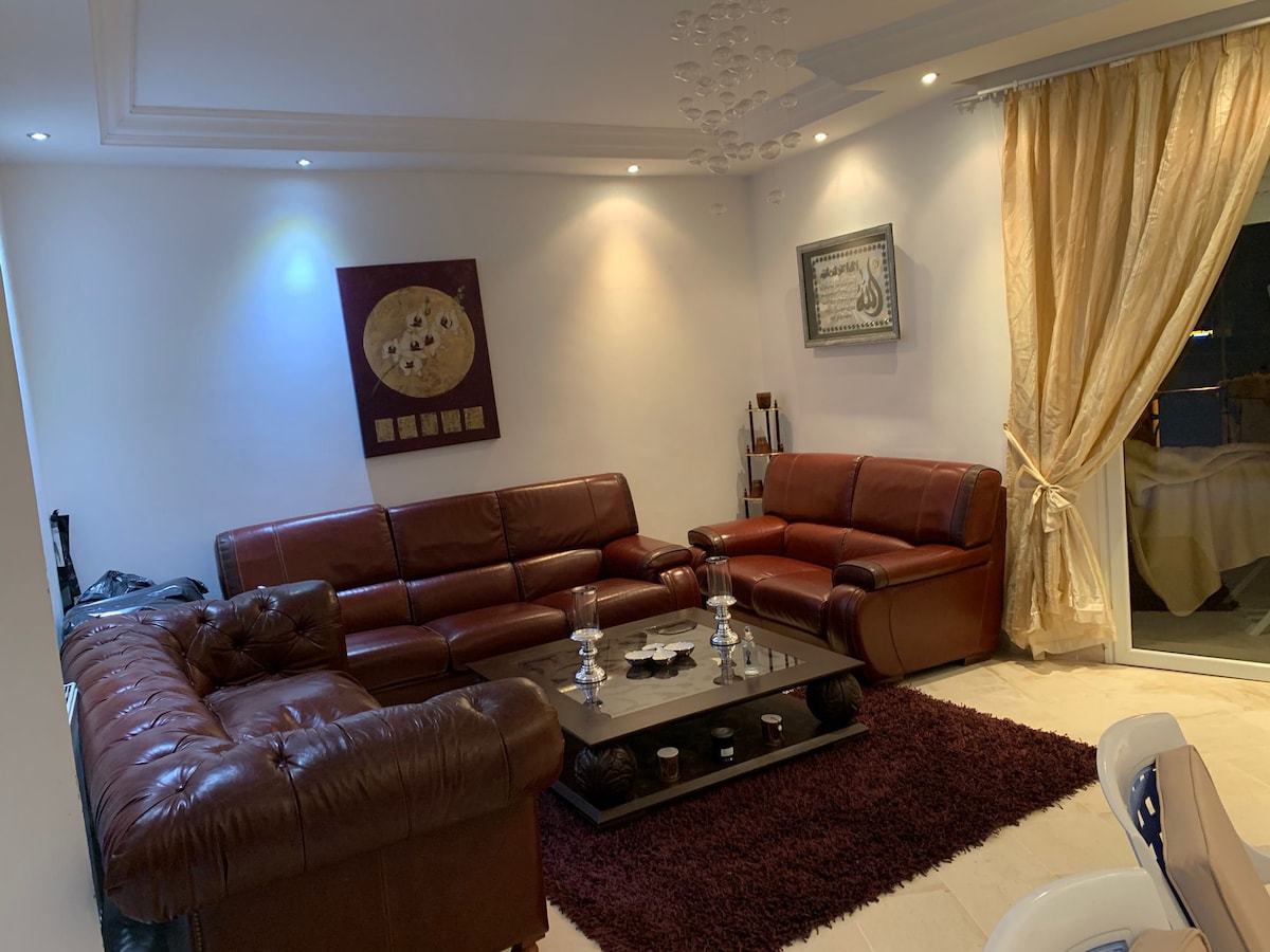 A living area features two dark leather sofas and a matching armchair arranged around a central coffee table. Soft lighting illuminates the space, complemented by a large area rug and floor-to-ceiling curtains. A decorative wall piece adds a subtle touch to the ambiance.
