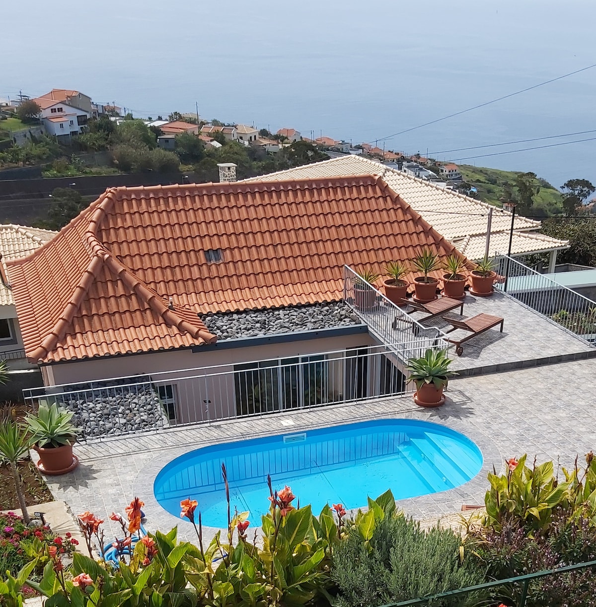 A spacious holiday home is situated atop a scenic hill, featuring a terracotta-tiled roof. Below, a serene pool is framed by potted plants, with colorful flowers enhancing the outdoor space. The vast ocean views create a tranquil backdrop for relaxation.
