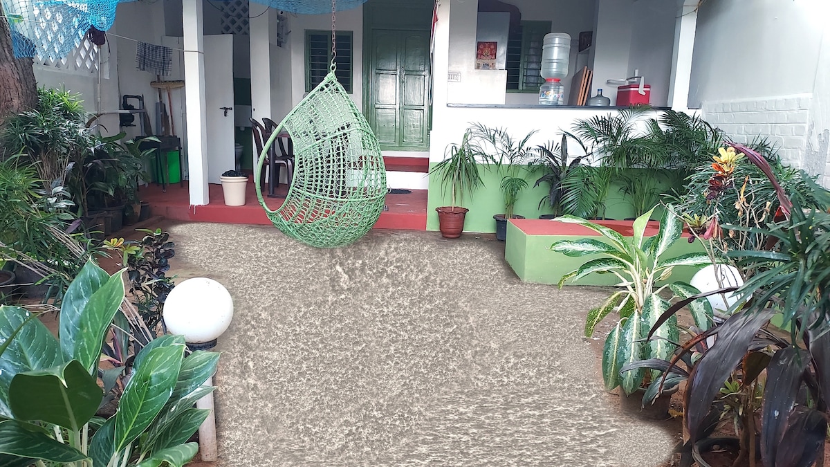 A vibrant garden area is adorned with lush greenery and tropical plants. A hanging chair adds a cozy touch to the space, with a view of a well-equipped kitchen area in the background. Soft lighting enhances the tranquil atmosphere.