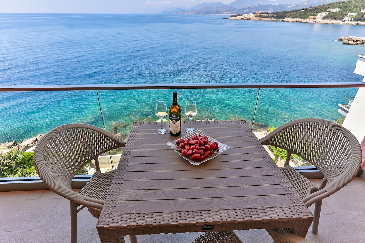 A balcony table is set for relaxation, featuring a plate of strawberries and two glasses beside a bottle of wine. The view of the clear blue sea and distant coastline creates a tranquil atmosphere. The glass railing offers unobstructed scenery of the vibrant water's edge.