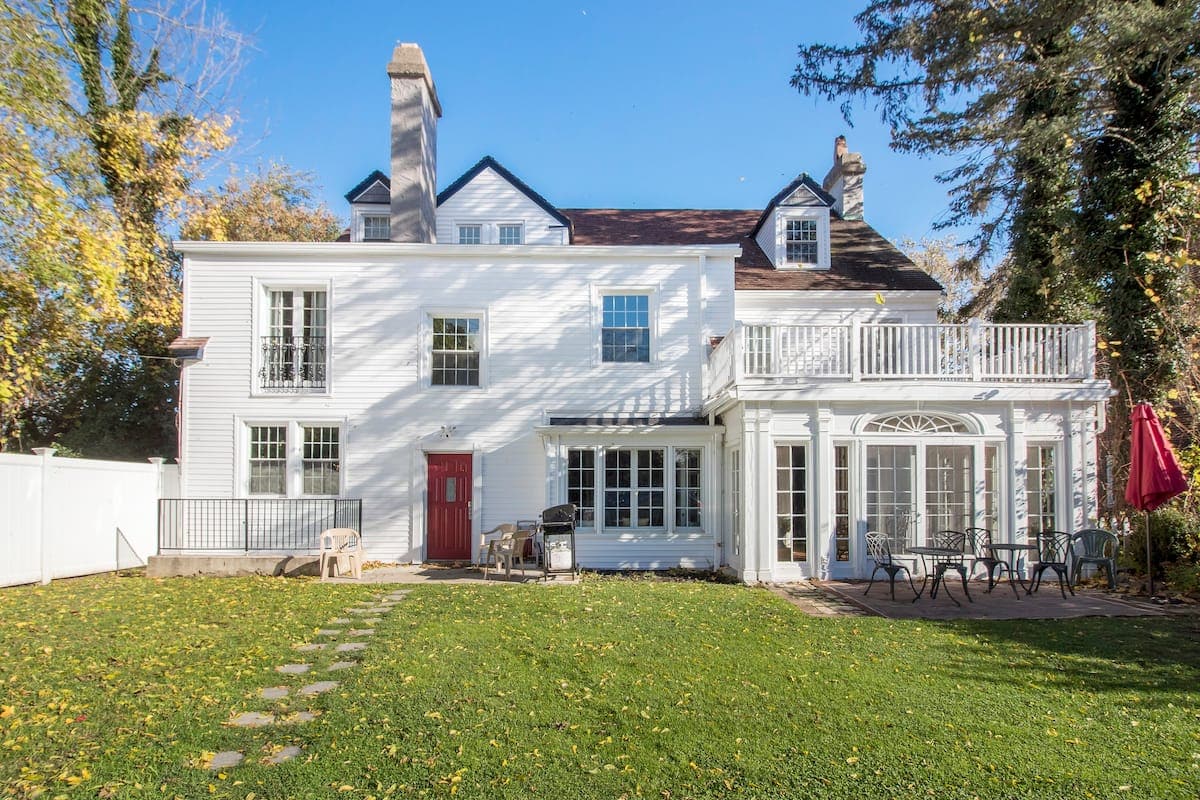 The spacious backyard features a well-maintained lawn with scattered autumn leaves. A patio area includes a dining table surrounded by chairs, with a red umbrella providing shade. The house, adorned with white siding, showcases large windows and a charming red door.