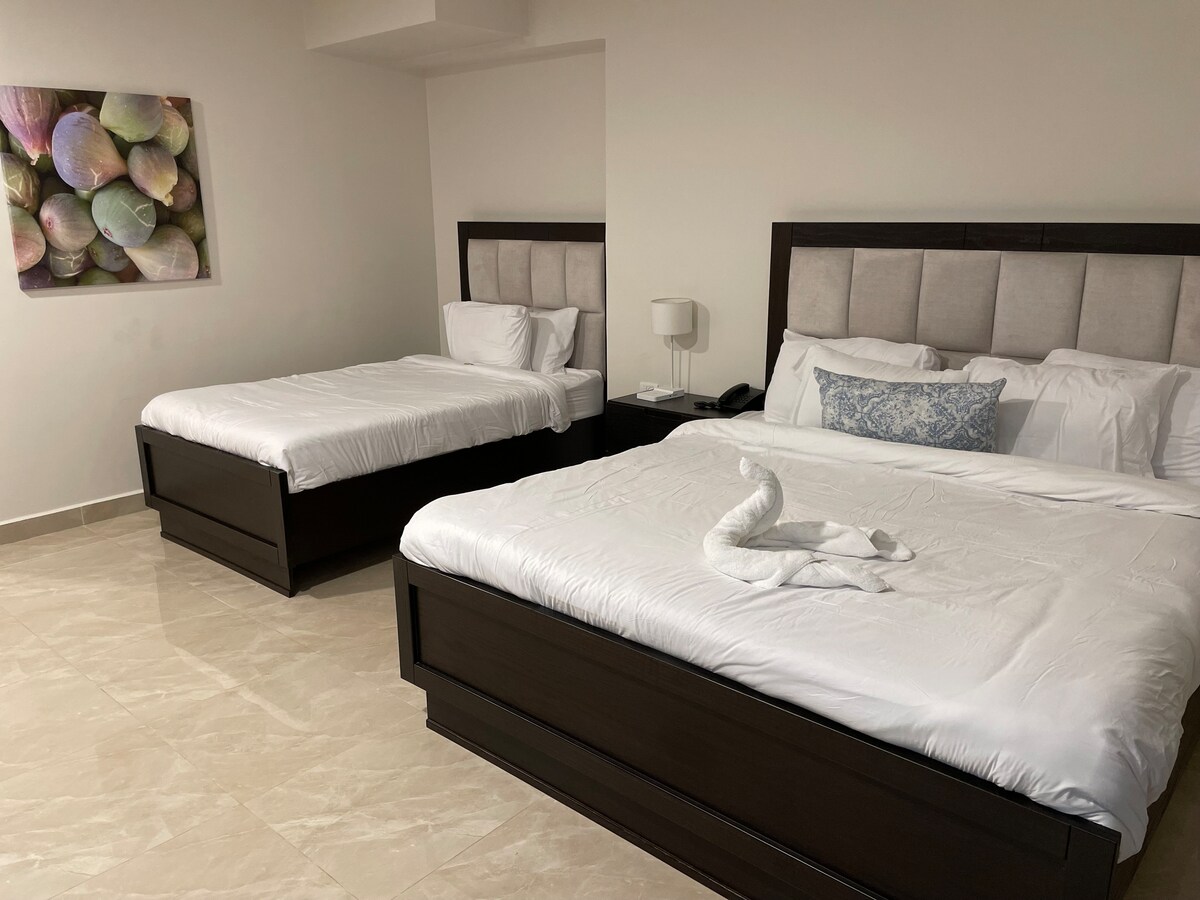 A spacious sleeping area features two large beds with white linens and decorative accents. A soft blue pillow adds a pop of color. A lamp and bedside table are positioned near one bed. Artwork depicting colorful shells adorns the wall, enhancing the room's aesthetic.