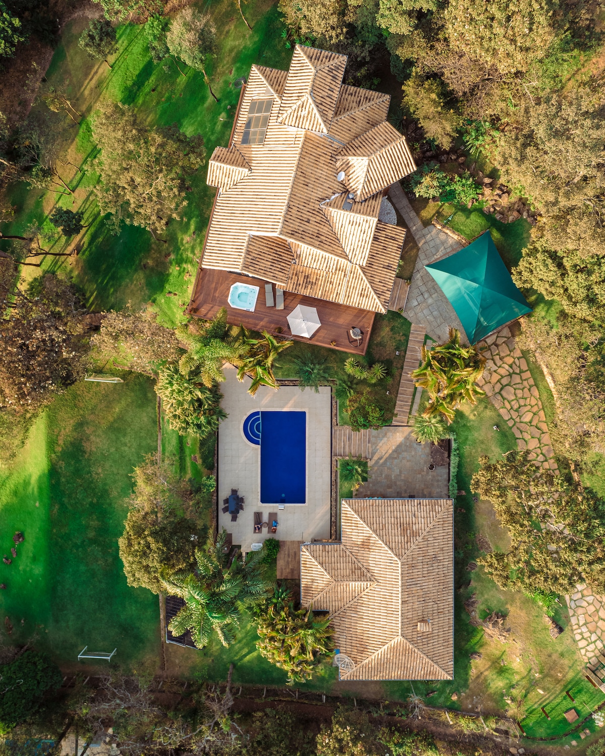An aerial view of a spacious property reveals a well-maintained lawn and a large swimming pool surrounded by lounge chairs. The layout includes multiple structures, including a covered gazebo and a hot tub area, all set within lush greenery.