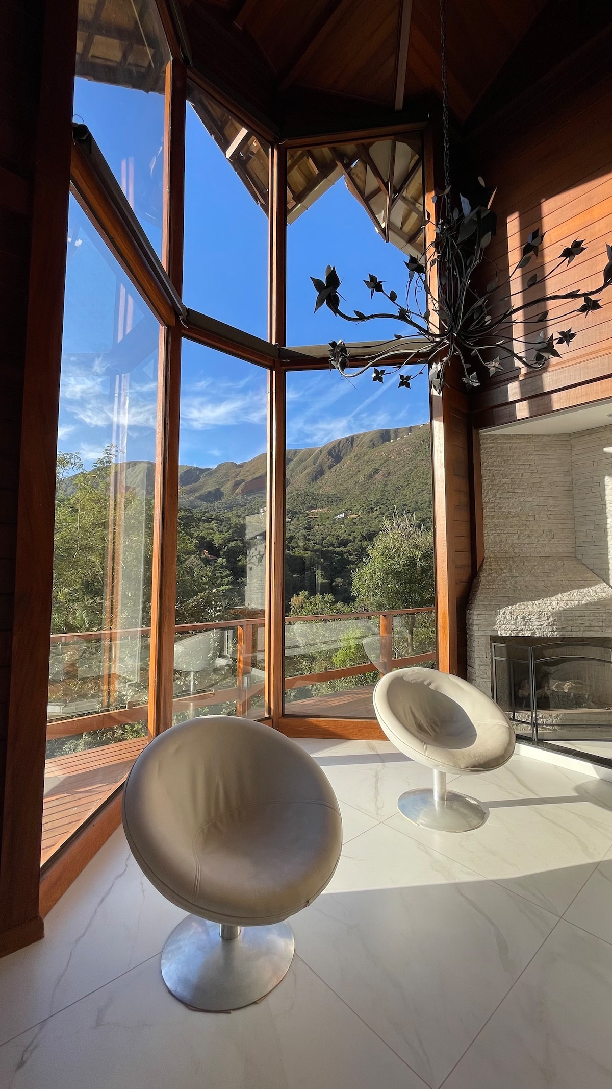 A bright seating area features two modern silver chairs positioned beside large floor-to-ceiling windows, offering panoramic views of the surrounding mountains. Natural light floods the space, highlighting the elegant chandelier above and the sleek marble flooring below.