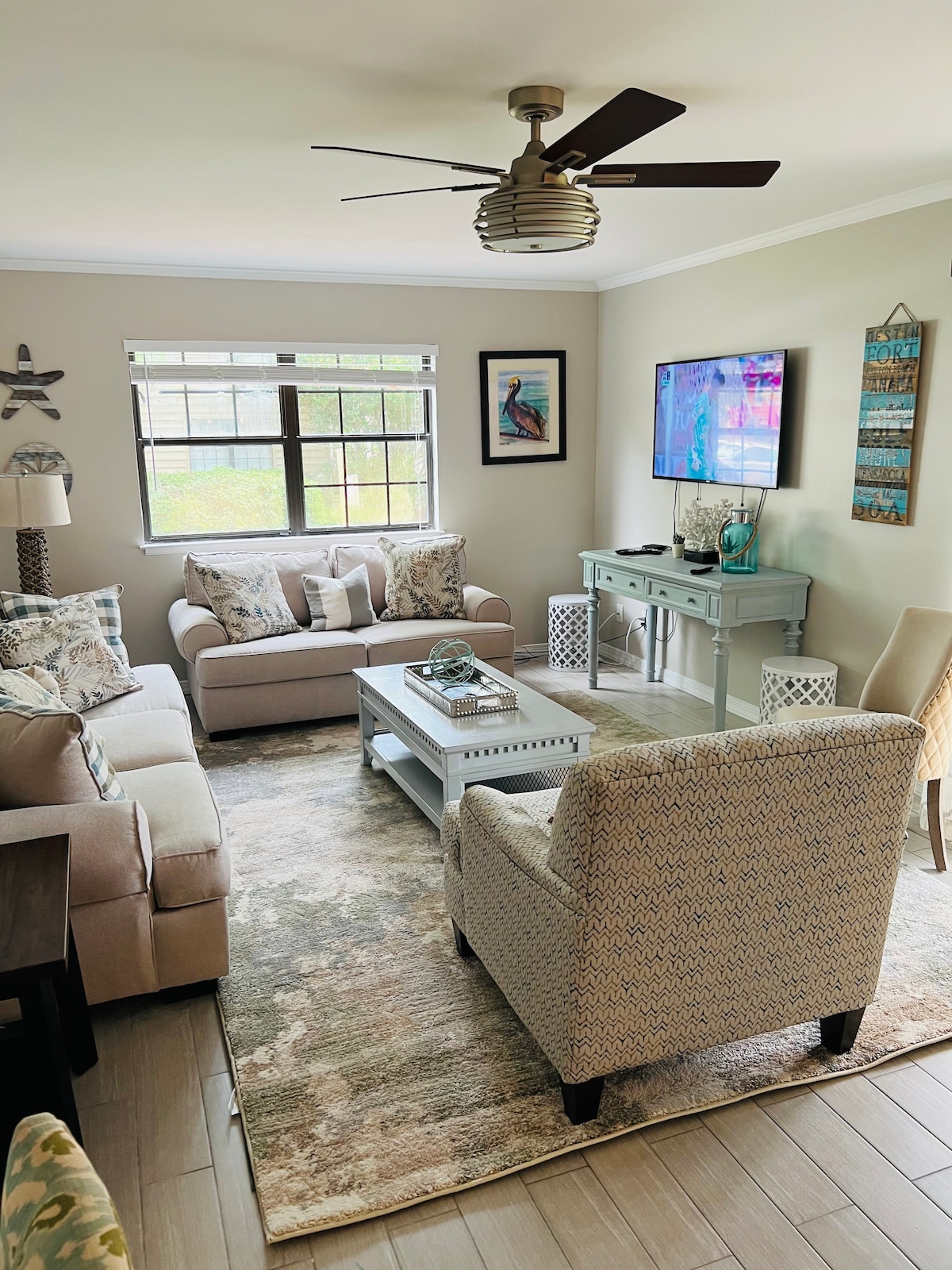 A cozy living area is presented with a light neutral color palette. Comfortable sofas are arranged around a coffee table, with decorative pillows enhancing their appeal. A ceiling fan provides airflow, while a television is mounted on the wall. Natural light filters through a large window.