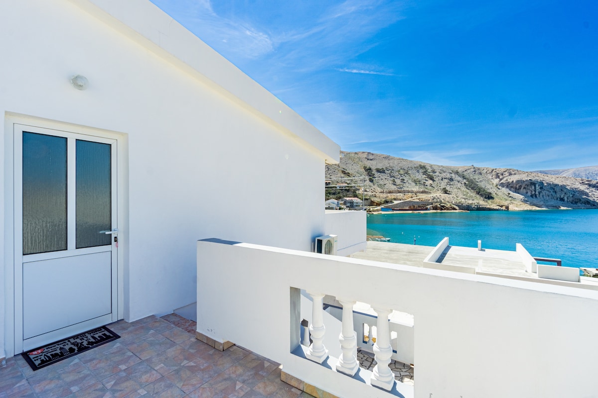 A bright entrance door is positioned beside a spacious balcony that overlooks a serene bay. The surrounding landscape features a mix of rocky hills and clear blue water, highlighting the outdoor tranquility. A simple welcome mat is placed near the door.