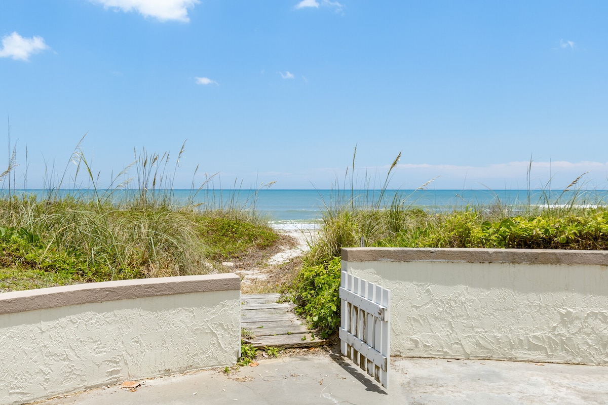 A sandy pathway leads from a white gate through tall grasses to the ocean. The blue sky meets the water's horizon, creating a serene atmosphere. The natural landscape invites relaxation and connection with the beach.
