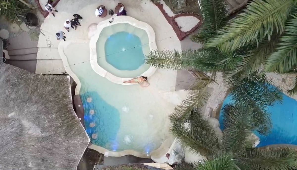 An aerial view captures multiple pools within a lush tropical setting, highlighting a circular jacuzzi surrounded by palm trees. Lounge chairs are positioned nearby, providing relaxation options for guests. The design features flowing curves and a harmonious integration with the natural surroundings.