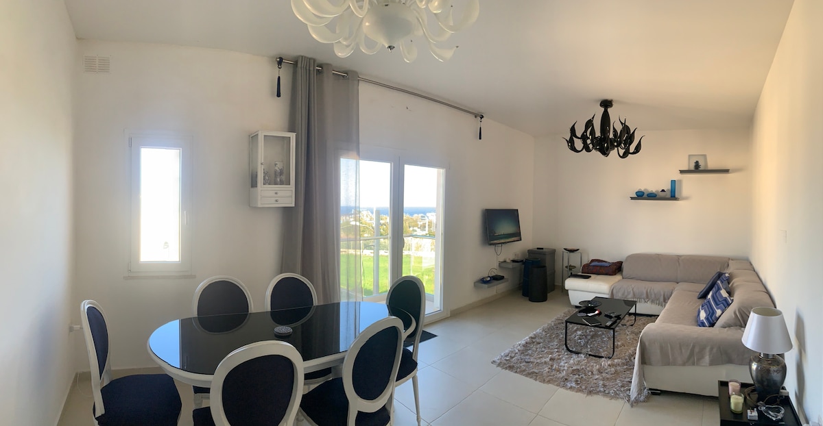 A spacious living area features a round dining table with dark chairs, adjacent to a light-colored sofa. Large windows provide a view of the sea and outdoor space. A decorative chandelier and minimalist decor enhance the bright and open ambiance.
