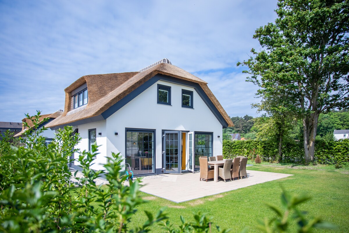 A modern thatched-roof villa is set against a clear sky, featuring large glass doors that lead to an outdoor dining area. A lush green lawn surrounds the property, complemented by well-maintained hedges and trees, contributing to a serene outdoor ambiance.