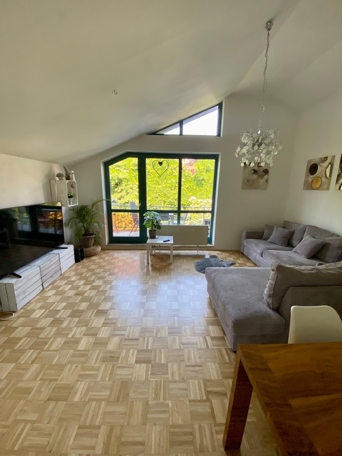 A bright living area features expansive windows allowing natural light to flood in. A gray sectional sofa is positioned near a contemporary TV stand, while a wooden dining table complements the space. Greenery is visible outside through the large glass panes, enhancing the room's ambiance.