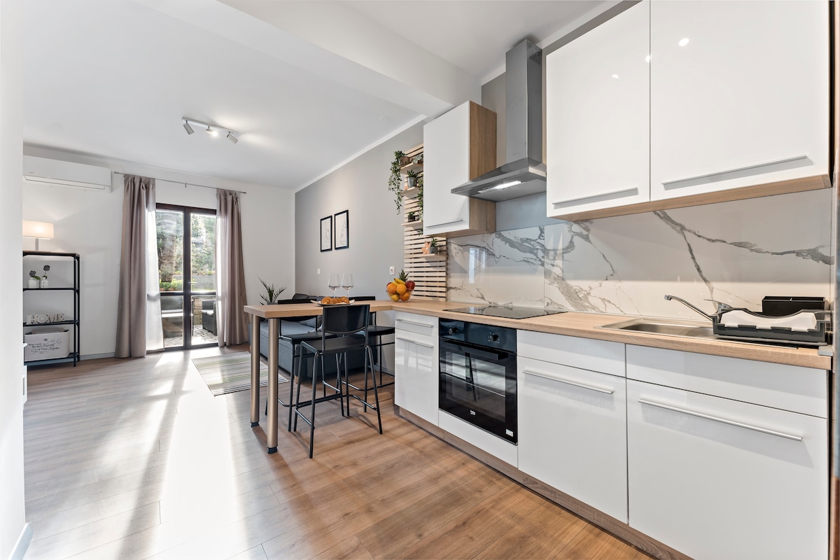 A bright kitchen area features modern cabinetry with a sleek countertop. Bar stools surround a small dining table, and natural light filters in through sliding glass doors leading to a terrace. Minimalist decor enhances the clean and organized layout.