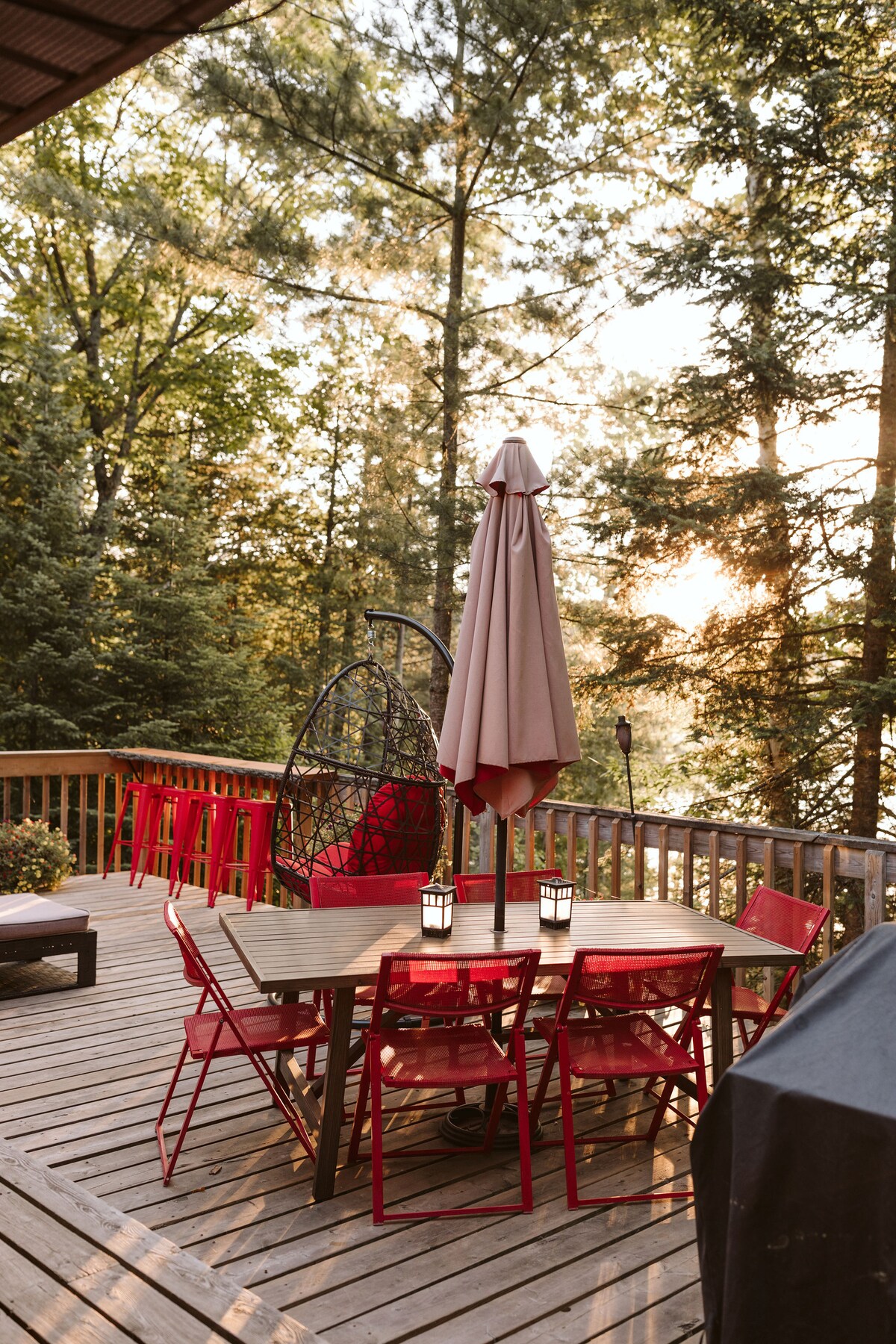 An outdoor deck area is displayed, featuring a dining table surrounded by red chairs. A large umbrella provides shade, while a hanging chair is positioned nearby. The space is framed by trees, creating a natural backdrop, and a grill is visible on one side.