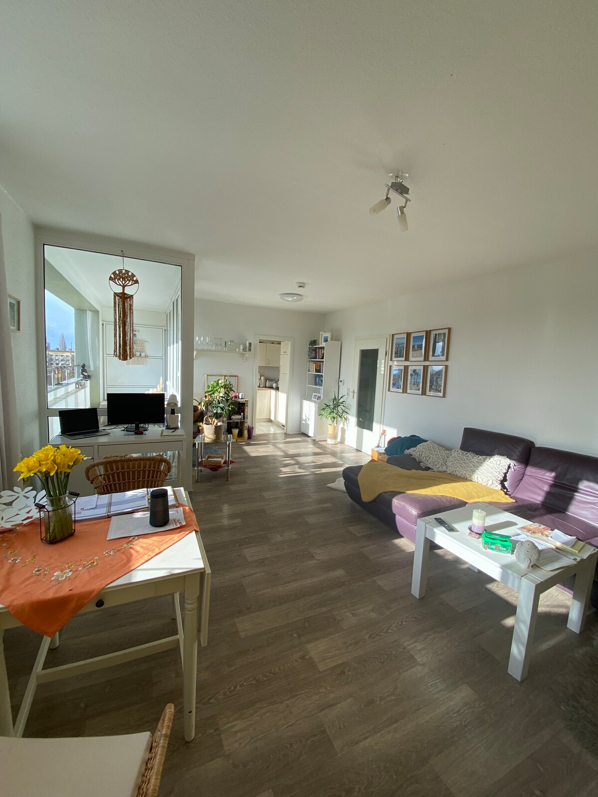 A bright living space is depicted, showcasing a comfortable seating arrangement with a purple sofa. A small dining table set with flowers and a computer is located near a mirror. Natural light fills the room, highlighting the modern decor and multiple framed pictures on the walls.