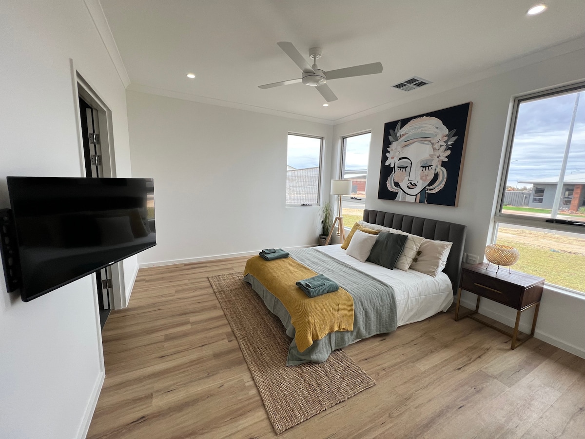 A spacious bedroom features a large bed dressed with light linens and a textured throw. Natural light floods in through two windows, and a wall-mounted smart TV is positioned across from the bed. A stylish side table complements the room, enhancing the modern design.