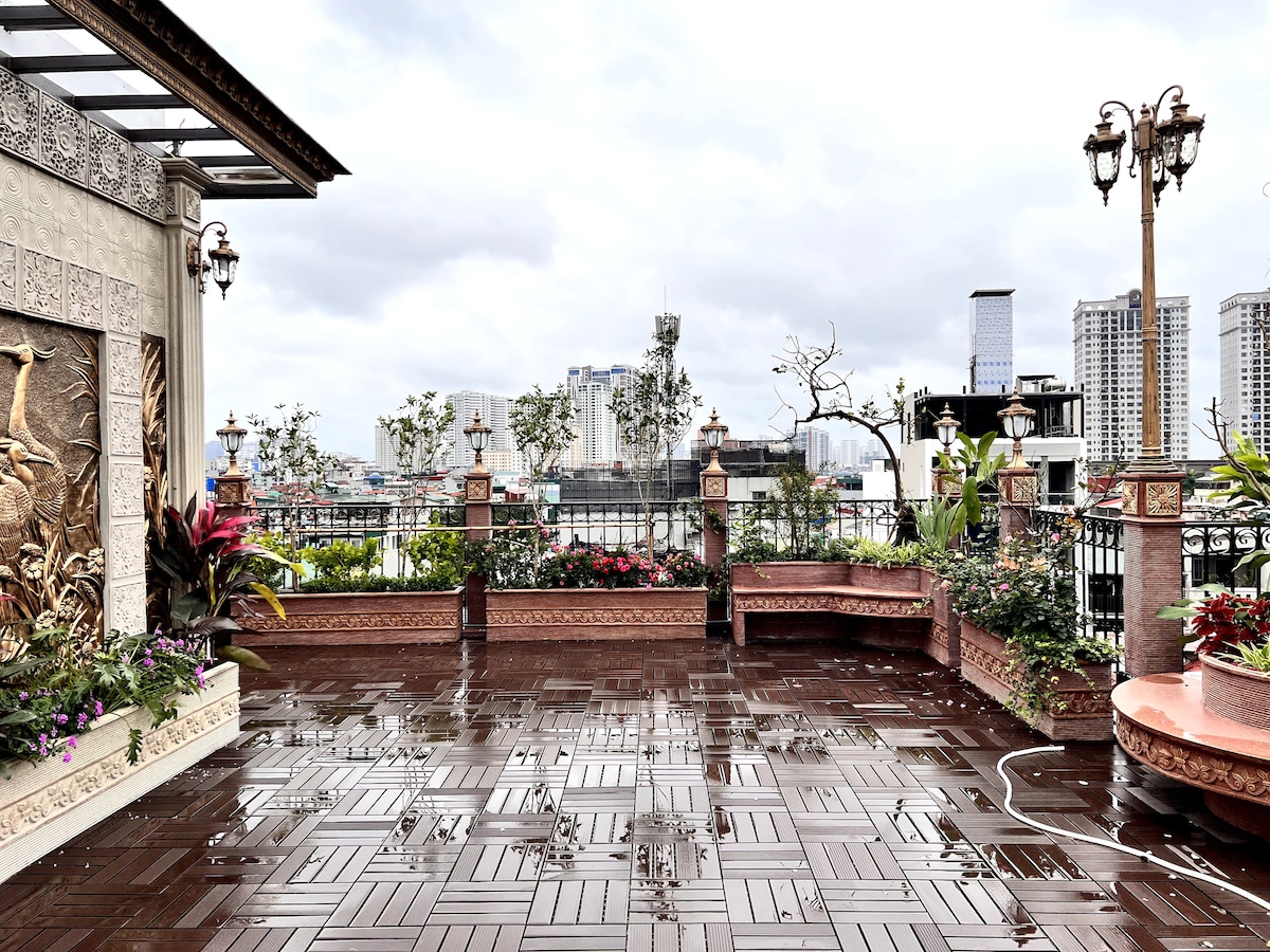 An outdoor rooftop terrace features an expansive tiled floor adorned with decorative planters filled with vibrant plants. Scenic views of the city skyline are visible in the background, complemented by artistic wall features and soft lighting fixtures along the edges.
