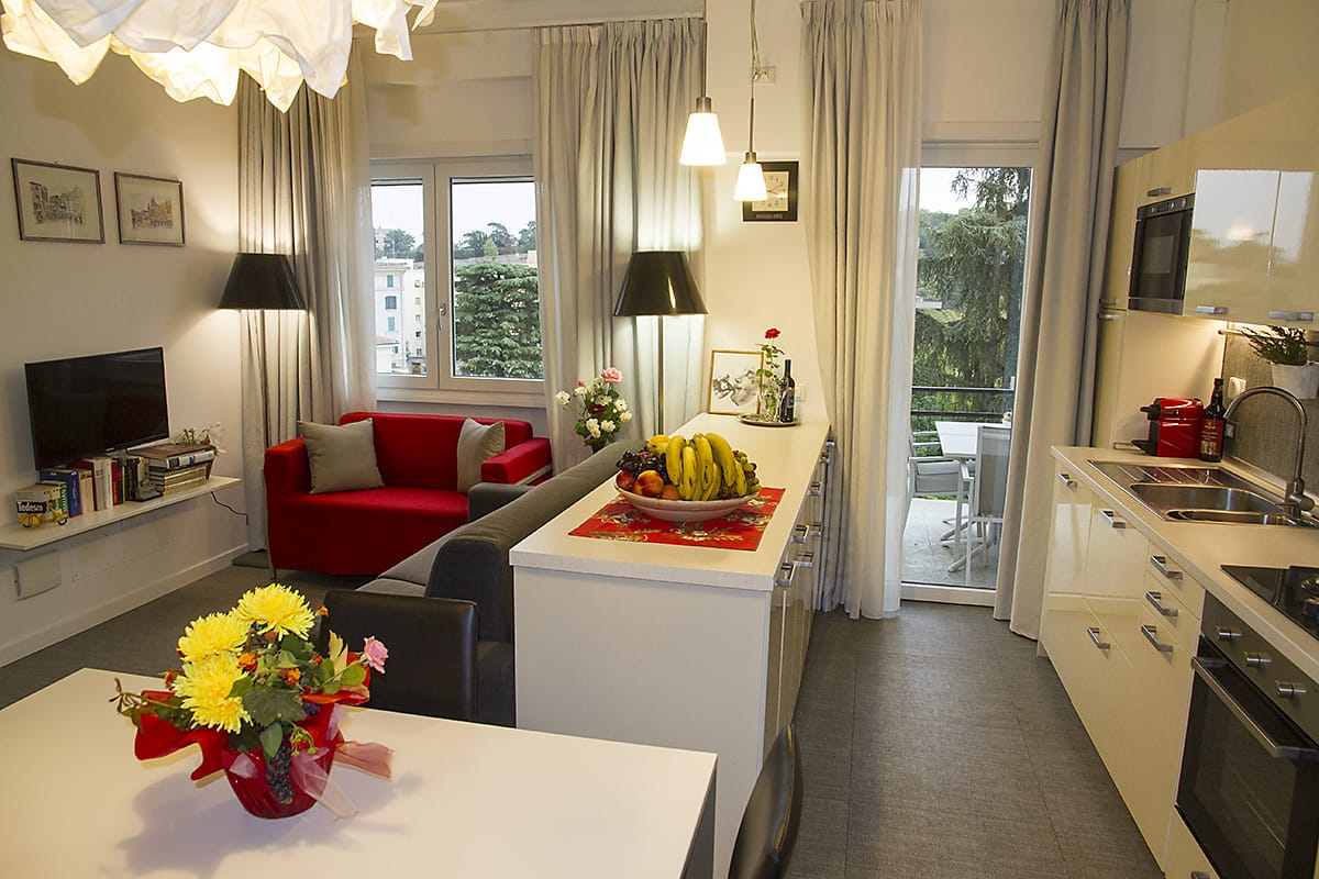 An open kitchen and living area is displayed, featuring a bright dining table adorned with a floral centerpiece. A cozy red sofa and a television are positioned near large windows that offer views of greenery. The kitchen includes modern appliances and ample counter space.