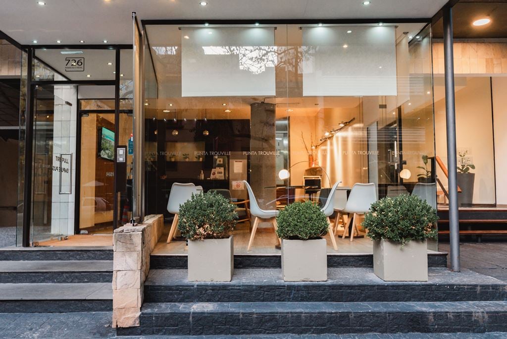 The entrance showcases a modern facade with large glass windows. Potted plants frame the entry steps, leading to a welcoming lobby area filled with stylish seating arrangements, enhancing the space's accessibility and comfort.