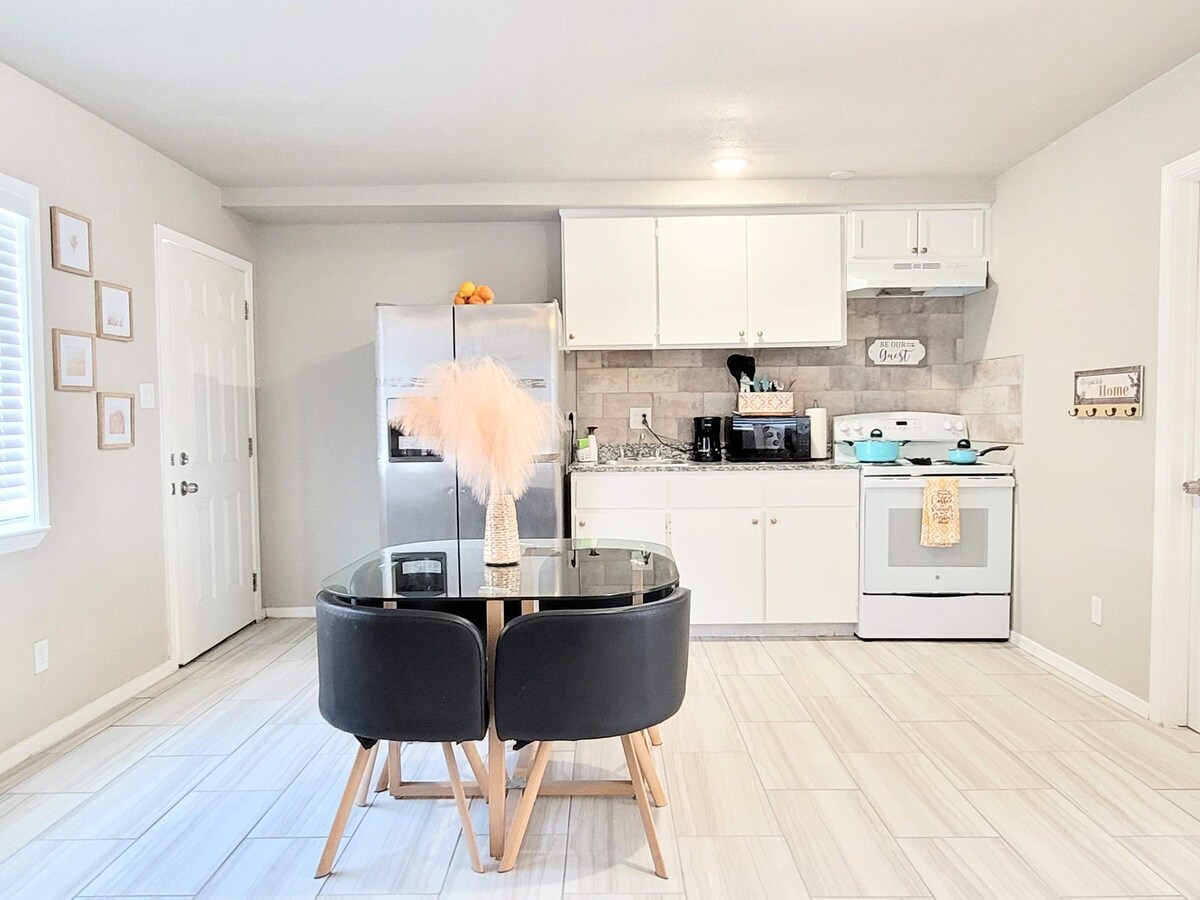 A modern kitchen area is featured with light tile flooring. A glass dining table is paired with black chairs. Stainless steel appliances include a refrigerator and stove, surrounded by white cabinetry and a neutral tile backsplash. Natural light enters through a nearby window.