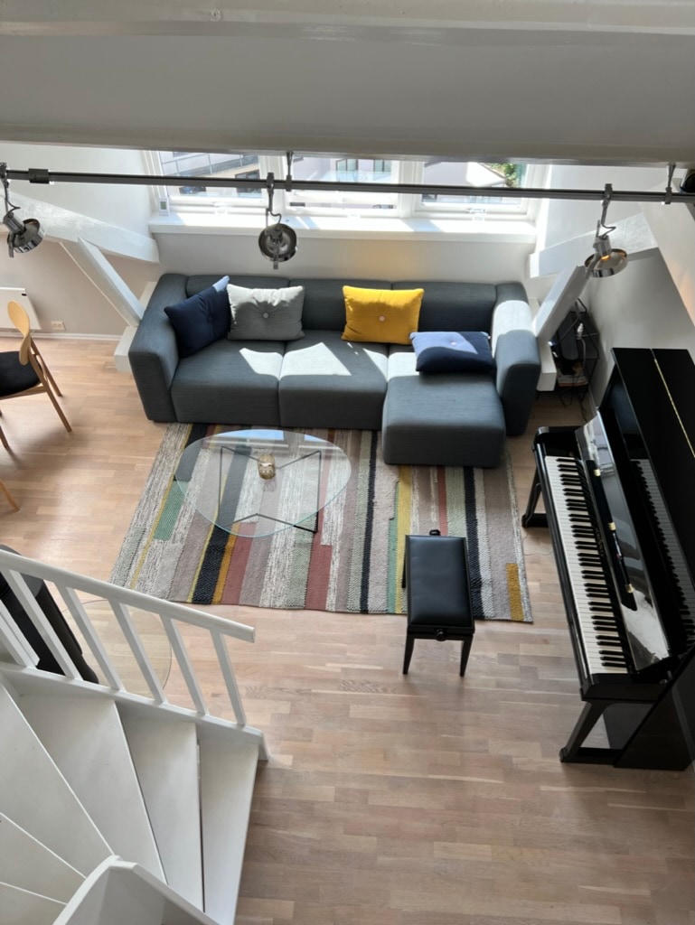 A bright and modern living area is visible, featuring a gray sectional sofa adorned with cushions in blue and yellow. A glass coffee table rests on a striped area rug. A piano is positioned nearby, with a black bench in front and large windows allowing ample natural light.