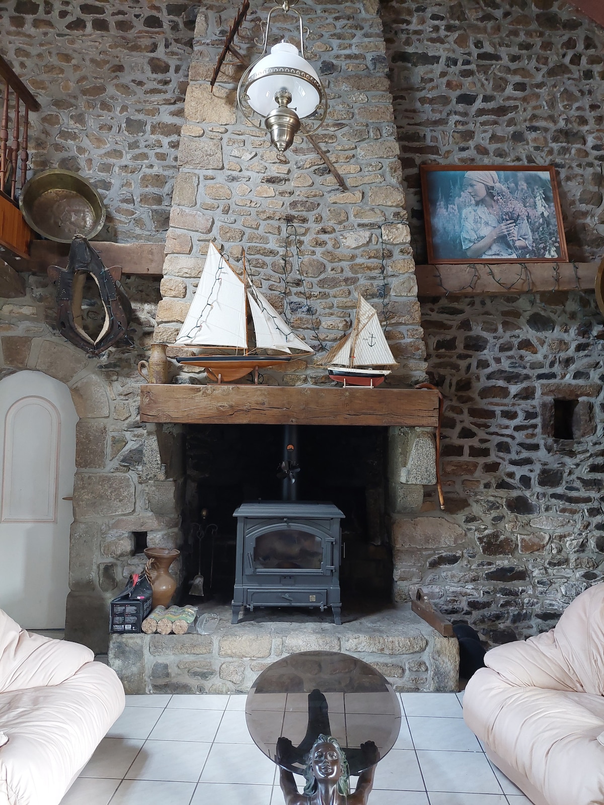 A large stone fireplace serves as a central feature in a cozy living area. Two soft, light-colored armchairs are positioned near a round glass coffee table. Nautical decor is displayed on the mantel, including small model sailboats and framed artwork, with textured stone walls providing a rustic backdrop.