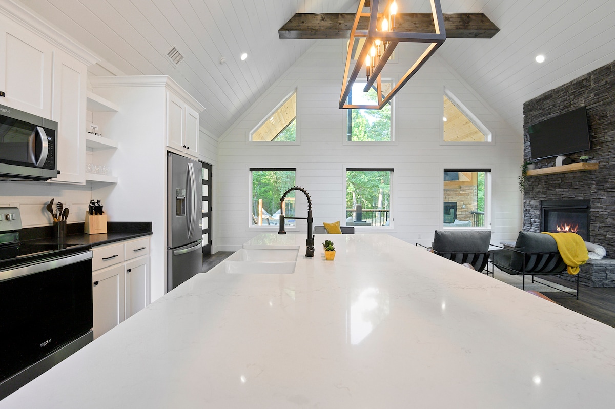 A modern kitchen boasts a spacious island with a marble countertop, complemented by sleek cabinetry. Large windows provide a view of nature, while a stone fireplace and stylish lighting fixtures add character to the inviting space.