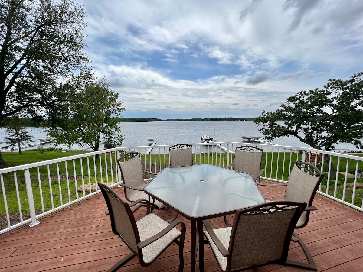 An outdoor dining area is set on a spacious wooden deck, featuring a glass-topped table surrounded by six chairs. The deck overlooks the lake, with expansive views of the water and tree-lined shores under a partly cloudy sky.