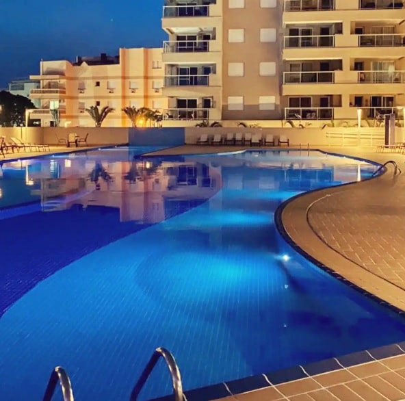 A large swimming pool is illuminated by soft blue lights, creating a serene ambiance. The pool features a curved design with clear water, surrounded by a spacious deck. Several lounge chairs are positioned nearby, offering a comfortable space for relaxation.