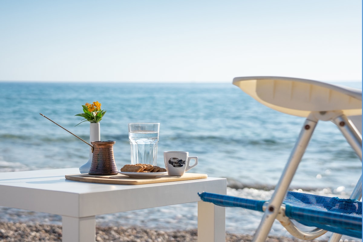 A serene outdoor setting with a small white table facing the sea. A glass of water, a coffee cup, and a plate with a pastry are arranged alongside a vase with fresh flowers. A lounge chair is positioned nearby, with clear blue water in the background.