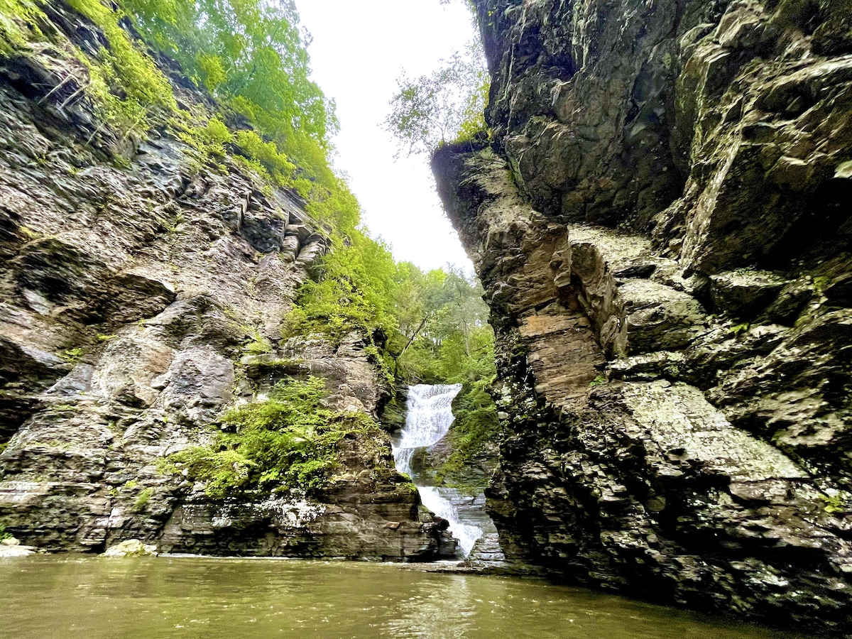 The image captures a cascading waterfall flowing between rocky cliffs, surrounded by lush greenery. Water fills a serene pool at the base, reflecting the natural beauty of the rugged landscape. The scene evokes a sense of tranquility and connection to nature.