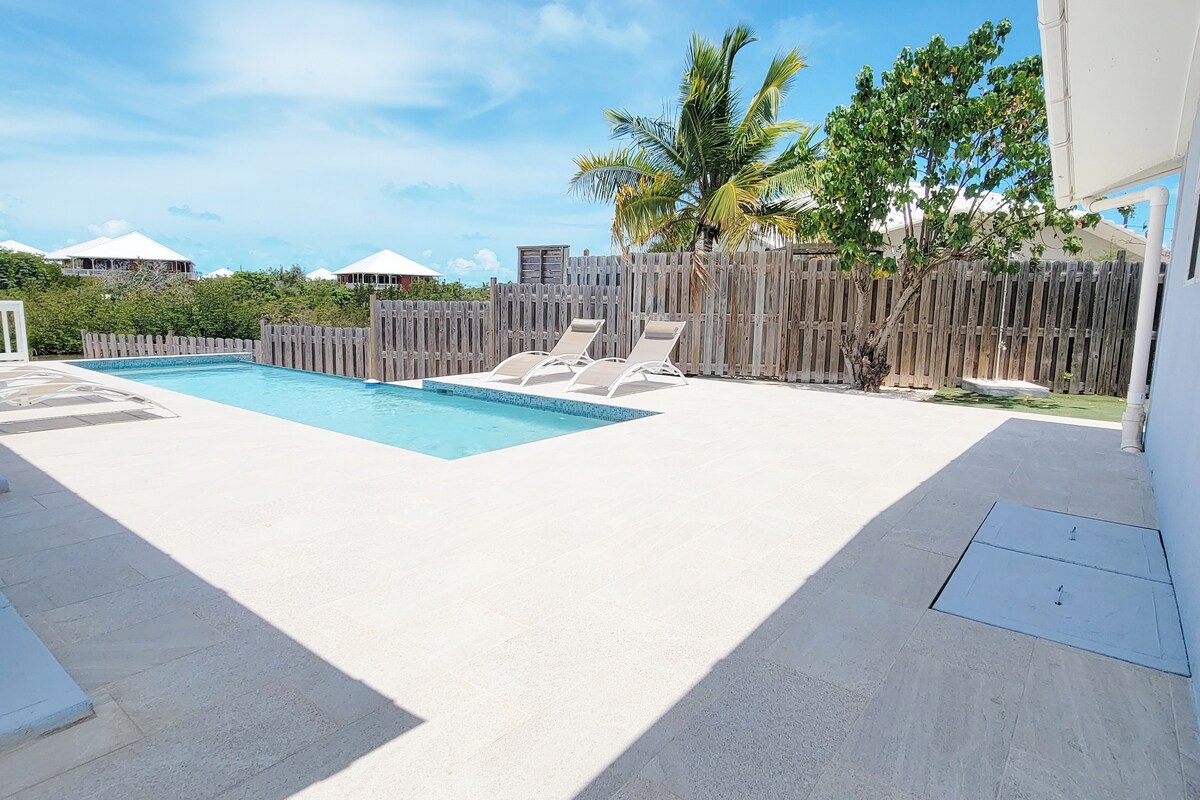 A tranquil pool area is surrounded by a smooth tiled deck featuring loungers for relaxation. Lush greenery and palms are visible in the background, along with a wooden fence that offers privacy. The clear blue water of the pool reflects the sunny sky.