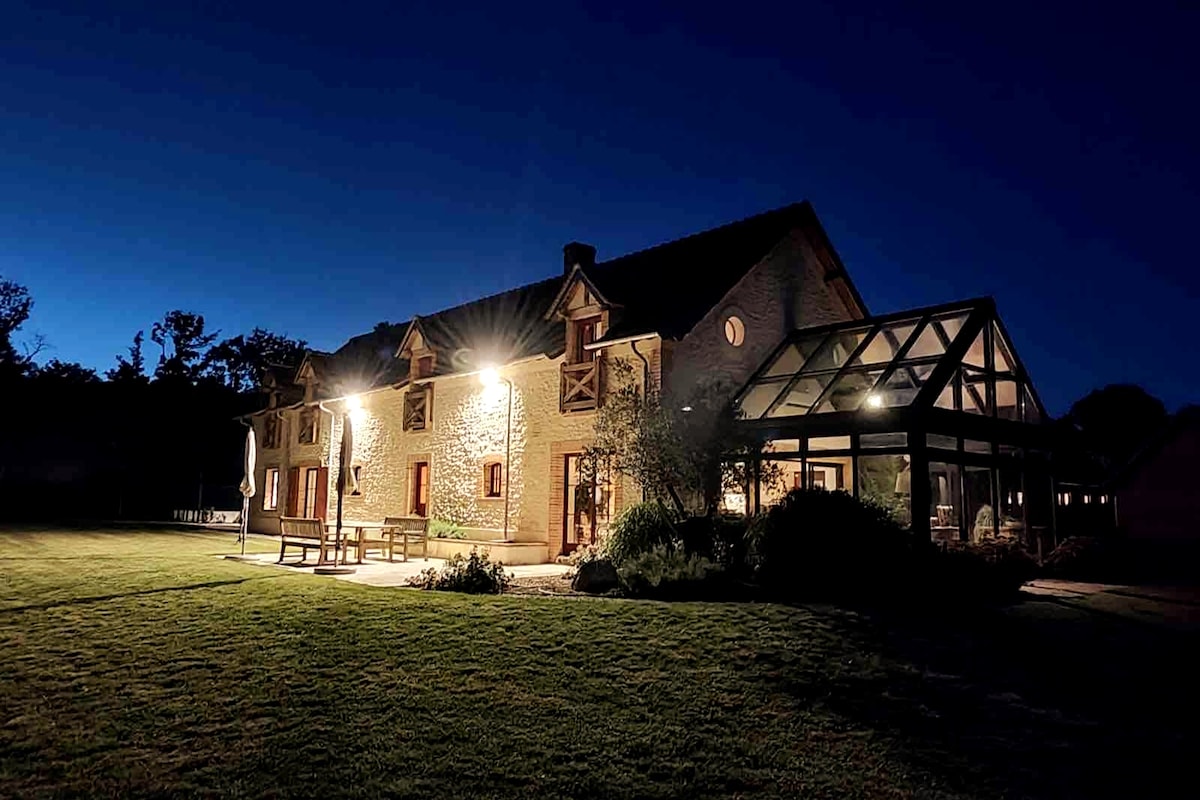 The stone house is illuminated under a twilight sky, showcasing its architectural details. A glass extension is visible, providing a seamless connection to the garden. Expansive green lawns surround the property, enhancing the serene outdoor setting.
