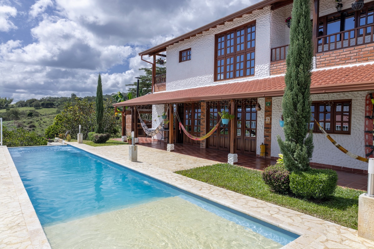 The exterior of the property showcases a spacious swimming pool with clear blue waters. Hammocks are placed under the covered terrace, adding a relaxed feel. The building features large windows and a brick facade, surrounded by lush greenery and trees, creating a serene landscape.