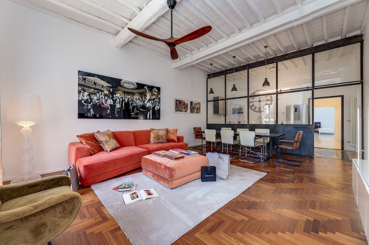 A spacious living area features a crimson sofa accompanied by decorative cushions, positioned near a unique coffee table. Artistic wall decor complements the room's aesthetic. A dining table with white chairs is visible, along with large windows allowing natural light to fill the space.
