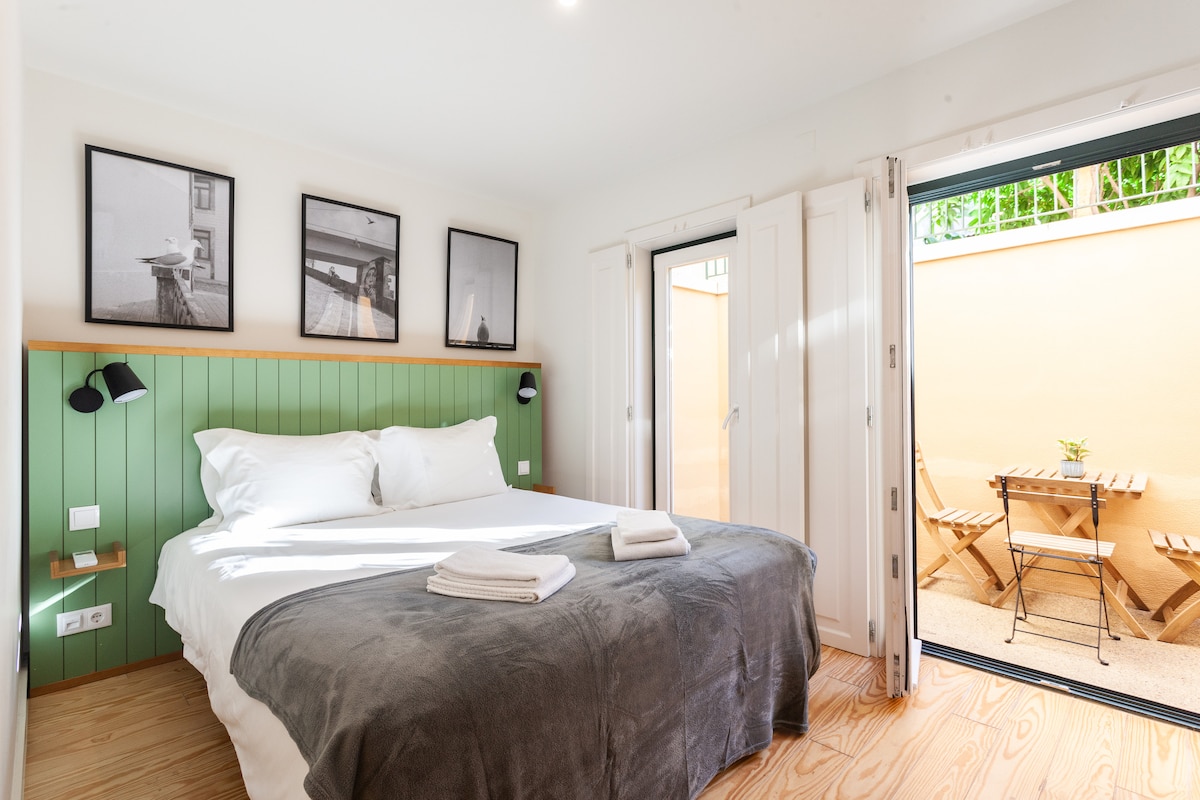 A bright bedroom features a spacious bed with a grey blanket and white linens. Black-and-white framed photographs hang on the wall, and sliding doors open to a private patio with a small table and chairs. The walls are accented in green, enhancing the calm ambiance.