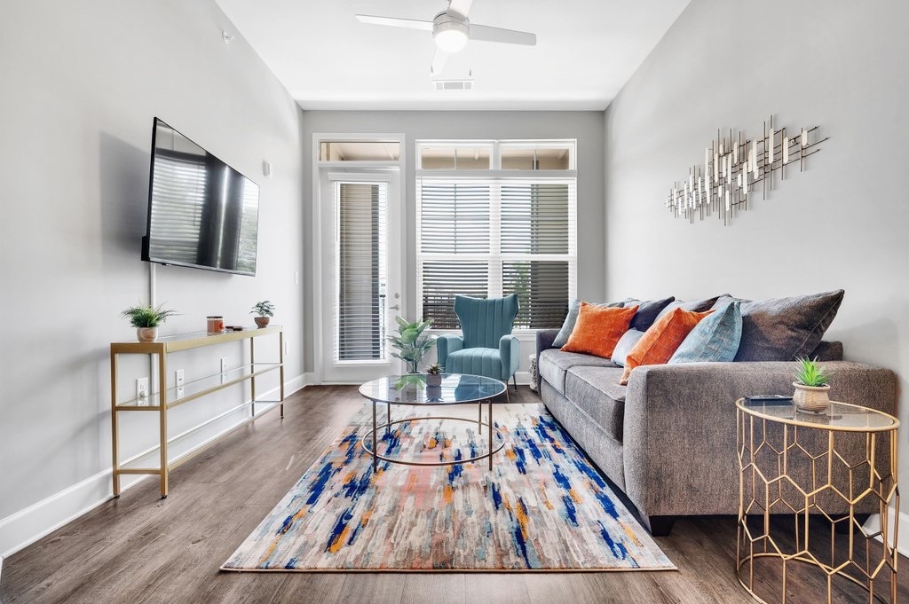 A cozy living area is furnished with a gray sofa adorned with colorful pillows and a vibrant area rug. A glass coffee table is centrally positioned, complemented by a stylish gold accent table. Large windows allow natural light to fill the space, enhancing its welcoming feel.