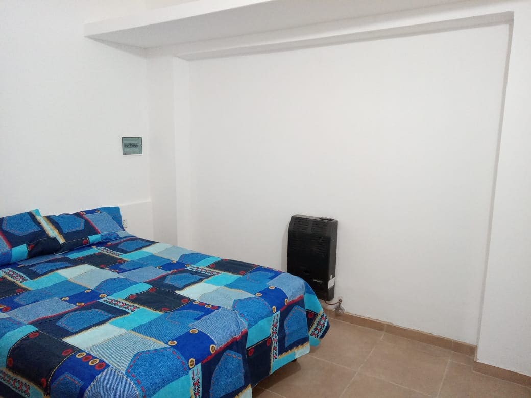 A sleeping area features a spacious bed covered with a blue patterned blanket. A white wall provides a simple backdrop, while a heater is positioned on the floor, adding functionality to the room. Soft natural light illuminates the space.