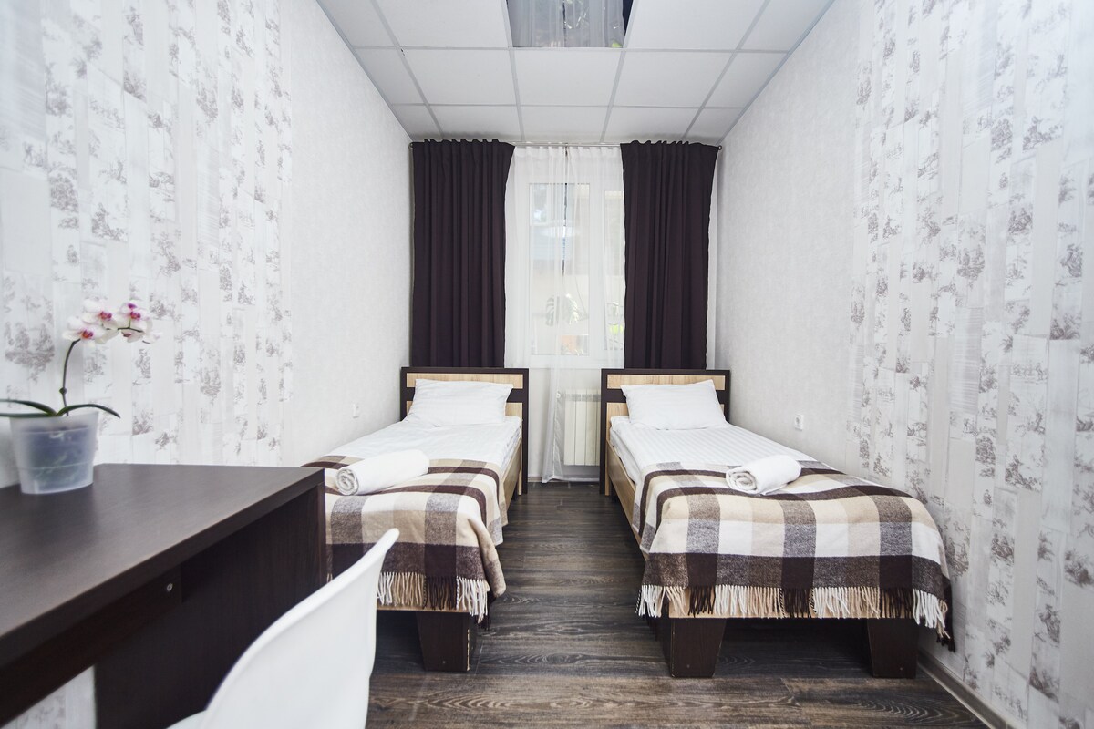 A well-lit room features two single beds with checkered bedding, positioned against light-colored walls adorned with subtle patterns. A small desk with a chair is visible, along with a window dressed in contrasting dark curtains, allowing natural light to fill the space.