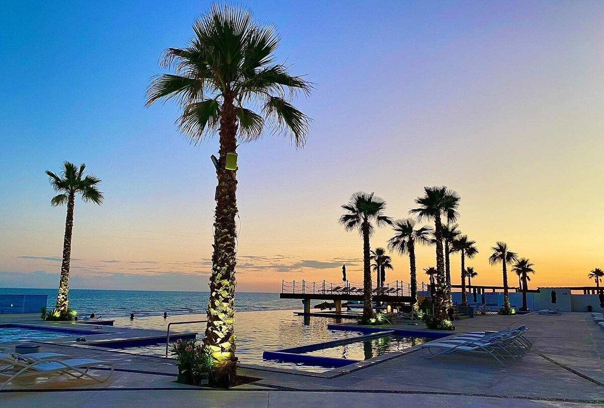 The outdoor area features a serene pool surrounded by several palm trees, which are illuminated softly. The tranquil ocean can be seen in the background under a pastel sky at dusk, creating a peaceful ambiance for relaxation.