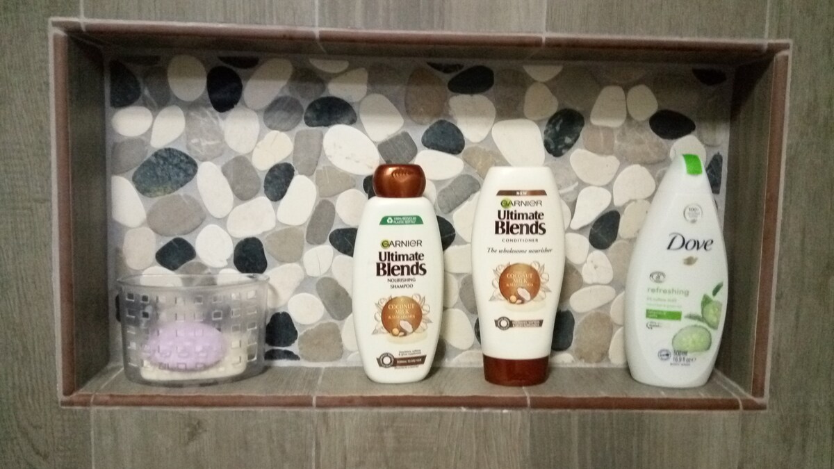 A bathroom niche displays a selection of personal care products, including two bottles of Garnier Ultimate Blends shampoo and conditioner, and one Dove body wash. A small, textured container holding a purple item is situated beside them, set against a backdrop of smooth, multicolored stones.