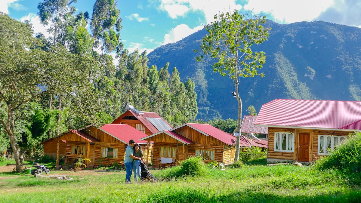 Several wooden cabins with red roofs are arranged on a grassy landscape. Lush trees surround the area, and a mountain range is visible in the background under a partly cloudy sky. A couple is seen together in the foreground, interacting with the environment.
