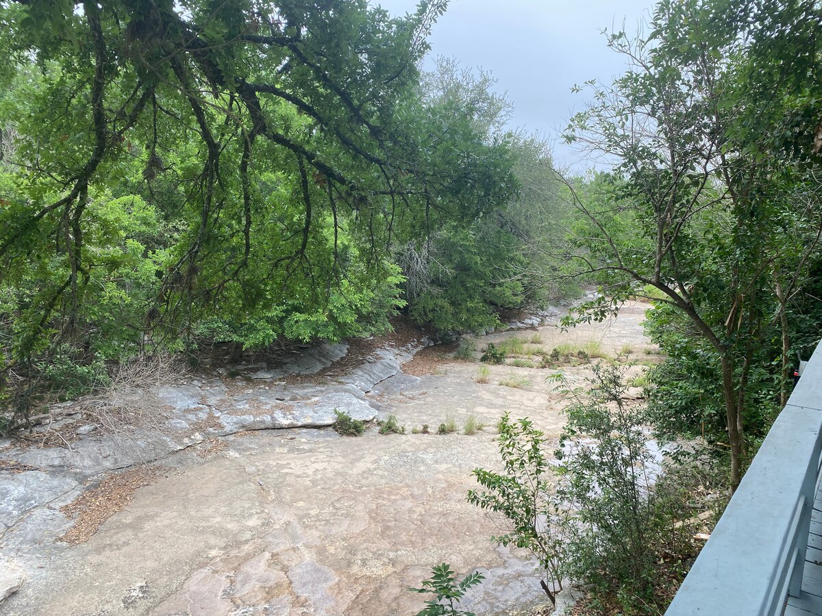 A serene view of a dry creek bed is framed by lush greenery. The rocky terrain displays muted earthy tones, while the surrounding trees create a natural canopy above, adding to the tranquility of the landscape.