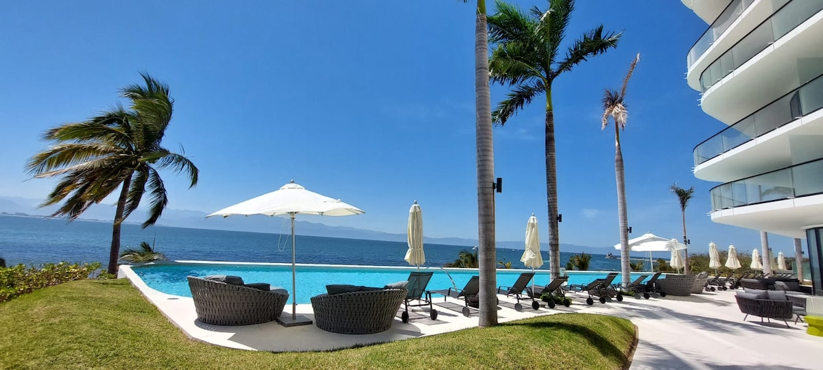 The image features a serene outdoor area beside the ocean, showcasing a large swimming pool. Sun loungers with umbrellas are arranged around the pool, and palm trees gently sway in the breeze. The clear blue sky complements the tranquil waters in the background.
